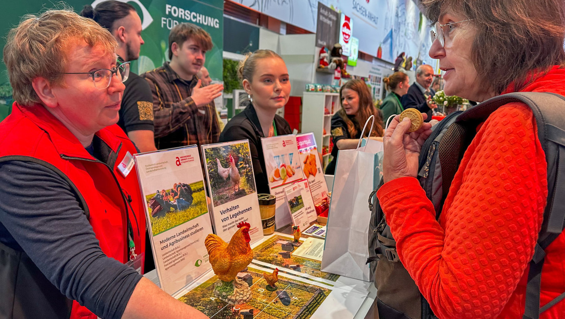 Zwei Frauen auf einer Messe stehen vor einem Infostand zur modernen Landwirtschaft und Geflügelhaltung. Eine hält Informationsbroschüren mit Bildern von Hühnern und Texten wie „Verhalten von Legehennen“, während die andere ein Stück Brot oder Gebäck in der Hand hält. Im Hintergrund sind weitere Besucher und Mitarbeiter zu sehen.