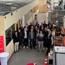 Gruppe von etwa 20 Personen steht lächelnd in einer industriellen Produktionshalle mit Maschinen und Rohrleitungen im Hintergrund. Einige halten Urkunden oder Zertifikate.