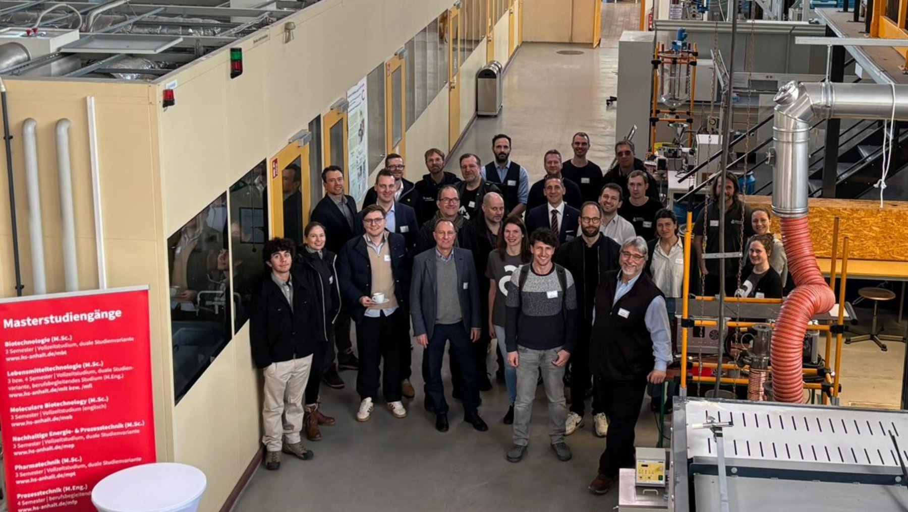 Gruppe von etwa 20 Personen steht lächelnd in einer industriellen Produktionshalle mit Maschinen und Rohrleitungen im Hintergrund. Einige halten Urkunden oder Zertifikate.